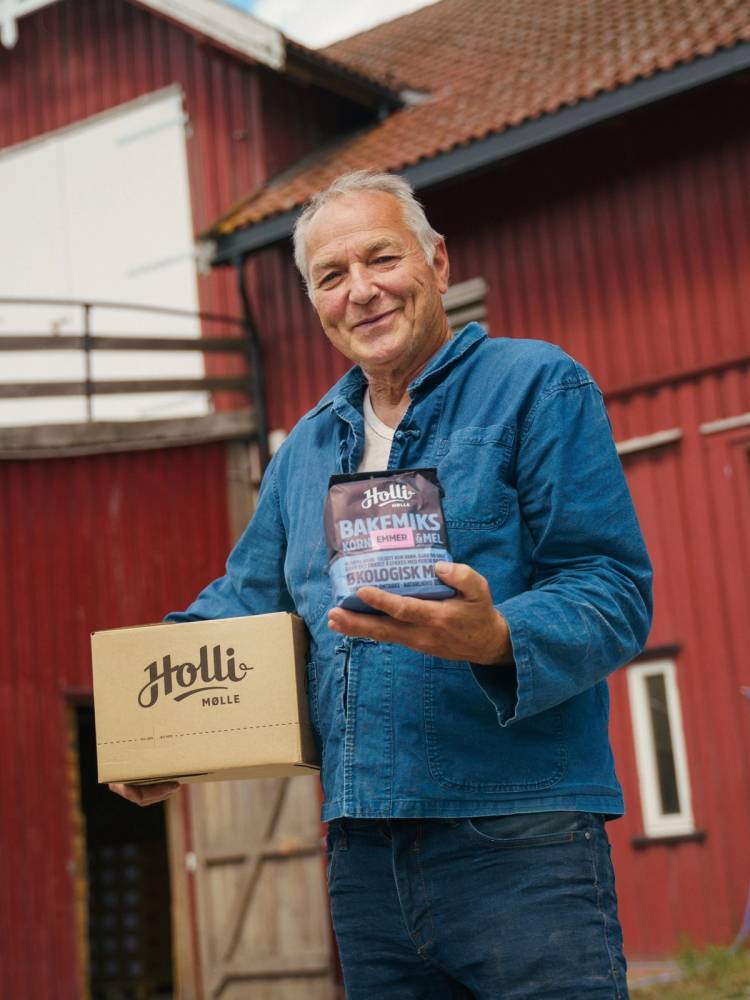 Person som holder en eske med Holli Mølle-brød og et pakke med bakeingredienser foran en rød låve.