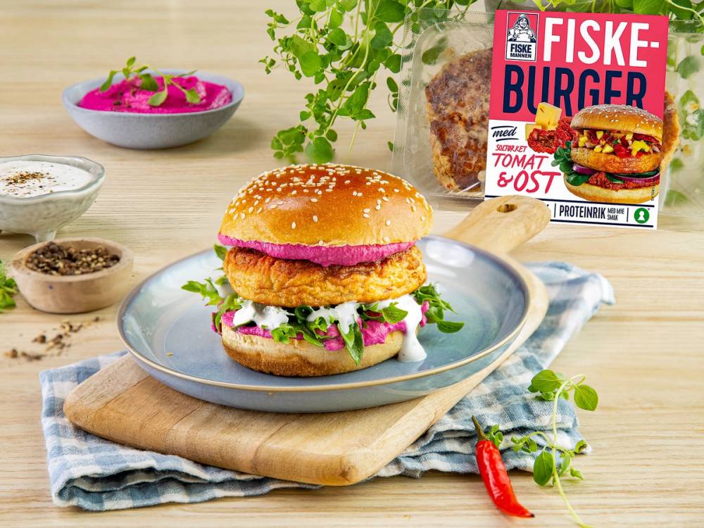 Fiskemannen-burger med fiskepinne, salat, tomat og ost på en tallerken, omgitt av krydder og urter på et kjøkkenbord.