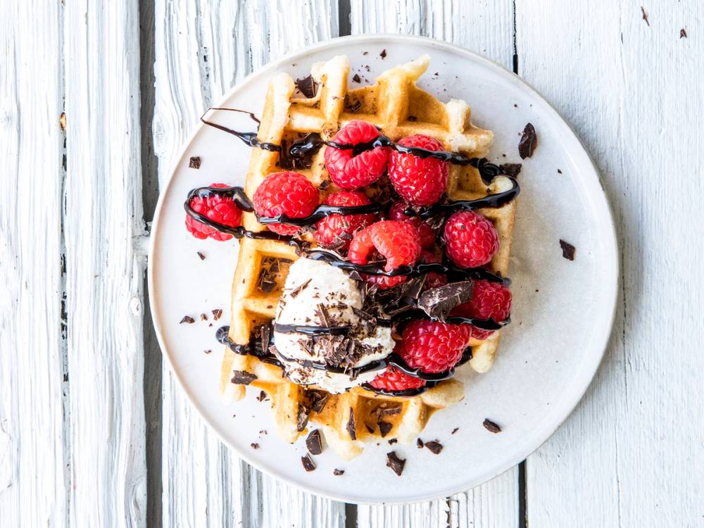 Belgisk vaffel toppet med friske bringebær, sjokoladesaus og et stykke sjokolade på en hvit tallerken, plassert på en hvit treflate.