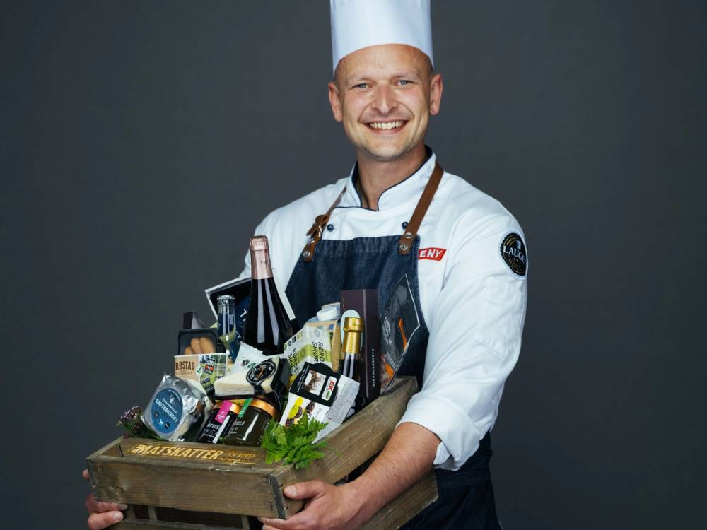 En person i kokkeuniform holder en stor eske med ulike matvarer og kjøkkenutstyr, mot en mørk bakgrunn.