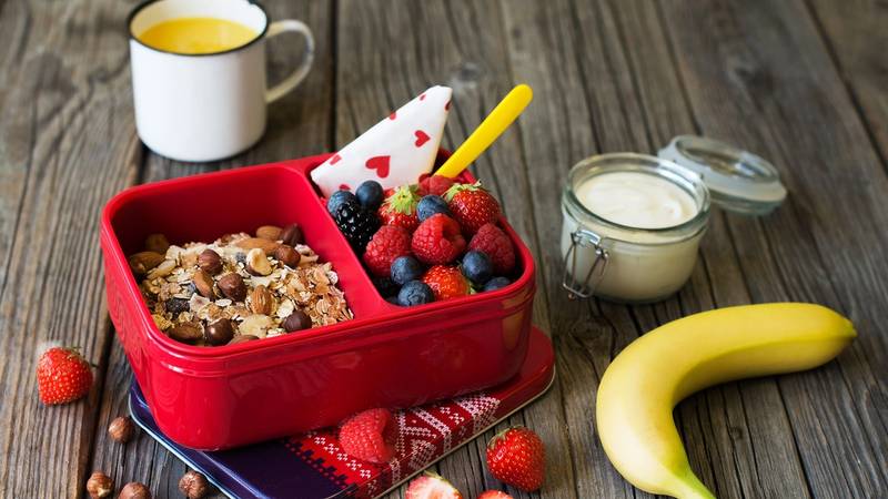 En matpakke med granola, bær og en banan på et bord, klar til å lages av barn.