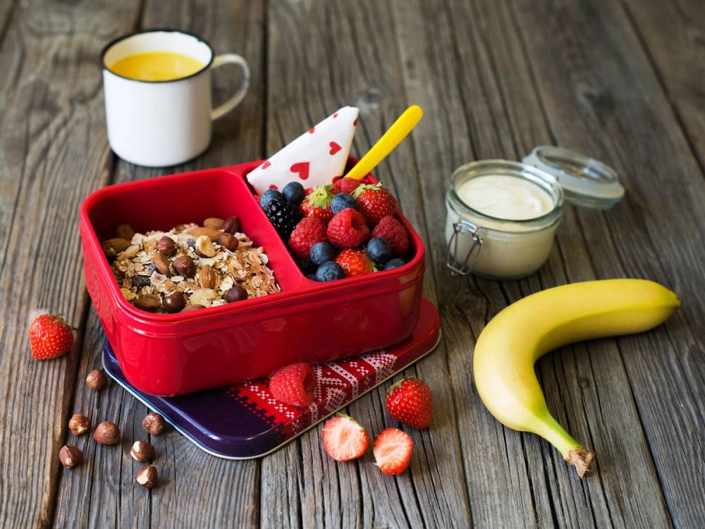 En matpakke med granola, bær og en banan på et bord, klar til å lages av barn.