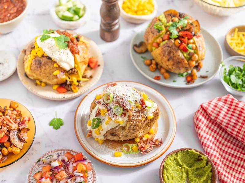 Bakt potet med tilbehør som rømme, ost og grønnsaker på en tallerken, omgitt av flere små skåler med tilbehør.