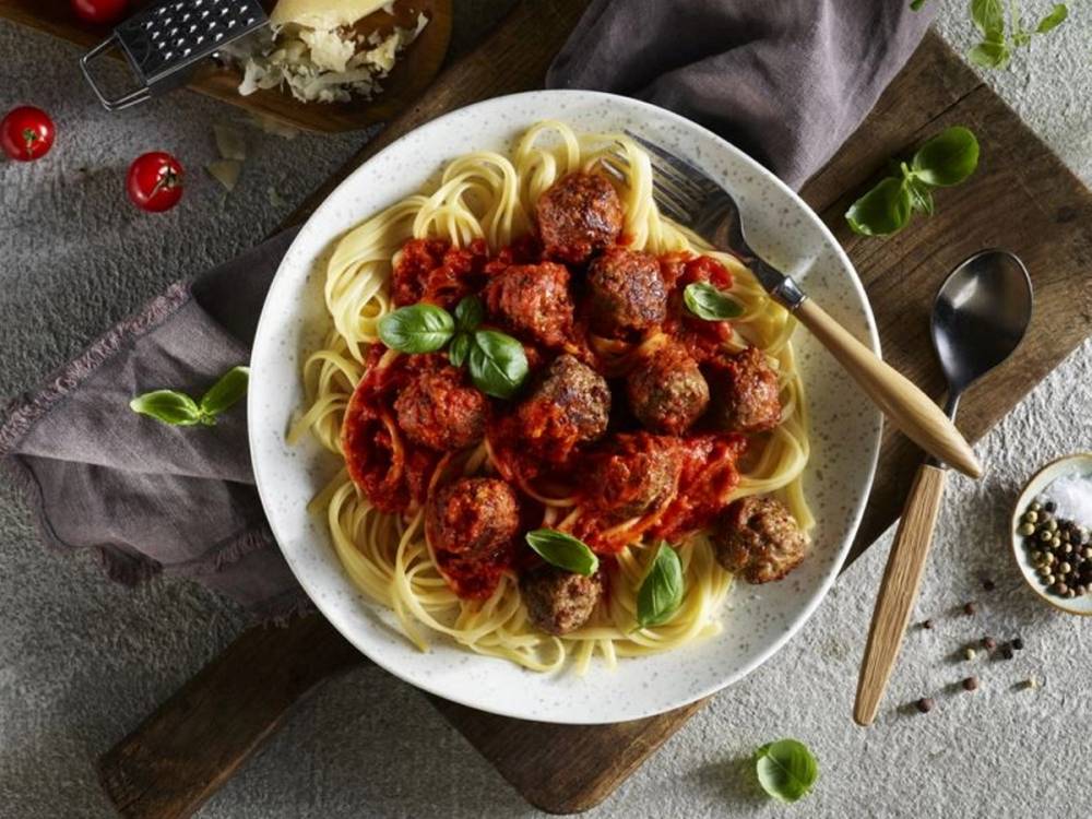 Kjøttboller i tomatsaus servert med spaghetti på en hvit tallerken, pyntet med basilikumblad.