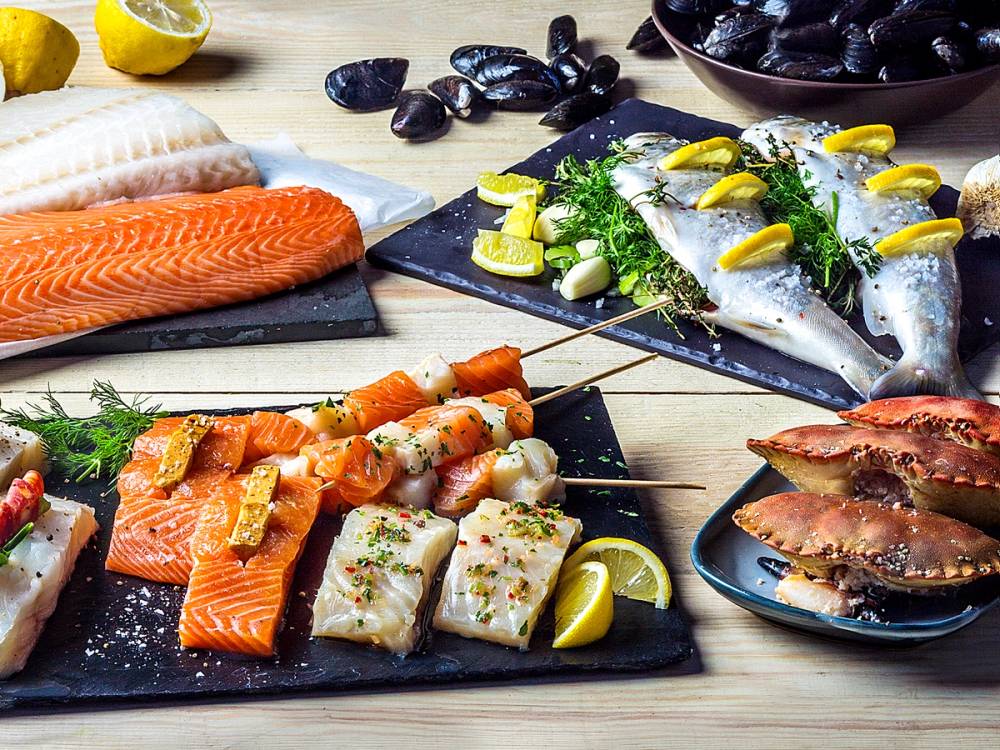Fiskeretter som laks, hvitfisk og skalldyr på grillen og på et serveringsfat, med sitronbåter og urter.