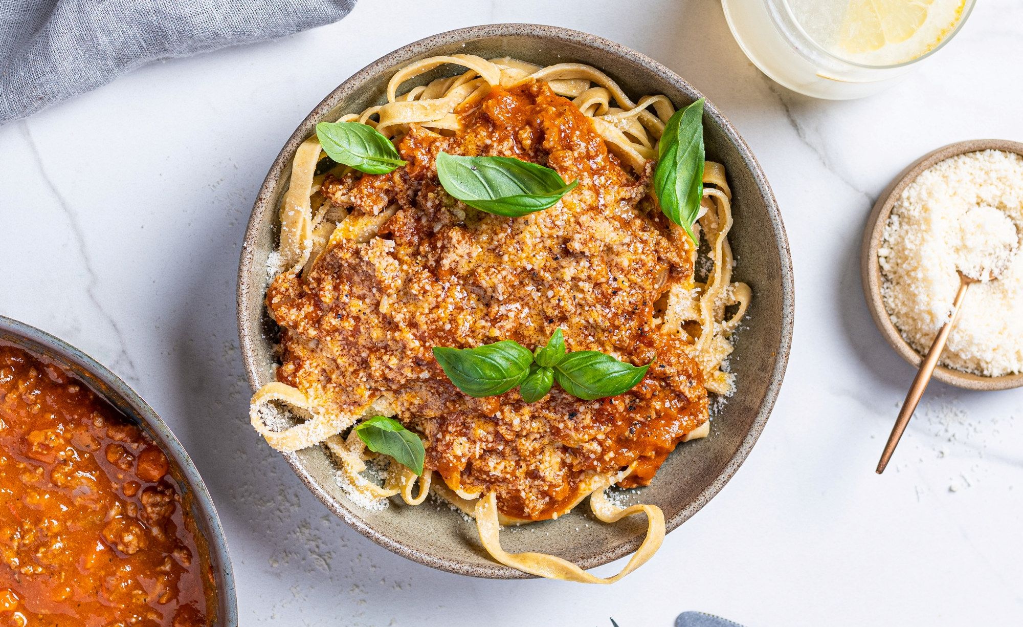 Pasta med kjøttsaus og ferske basilikumblader på toppen, servert i en svart skål med revet ost og en glass melk i bakgrunnen.