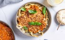 Pasta med kjøttsaus og ferske basilikumblader på toppen, servert i en svart skål med revet ost og en glass melk i bakgrunnen.