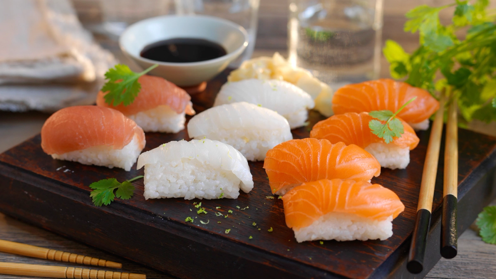 Sushi med laks og hvit ris på en svart serveringsplate med soyasaus og grønne urter i bakgrunnen