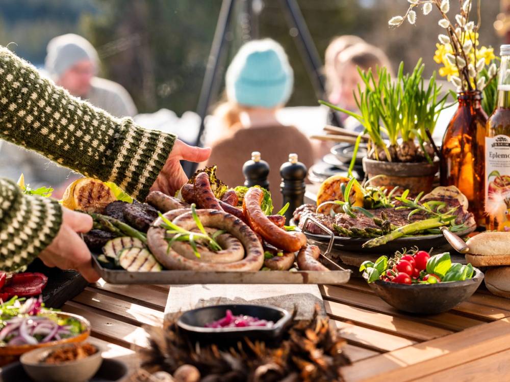 Utebord med påskemat som pølser, salater og grønnsaker, med personer i bakgrunnen som nyter solen.