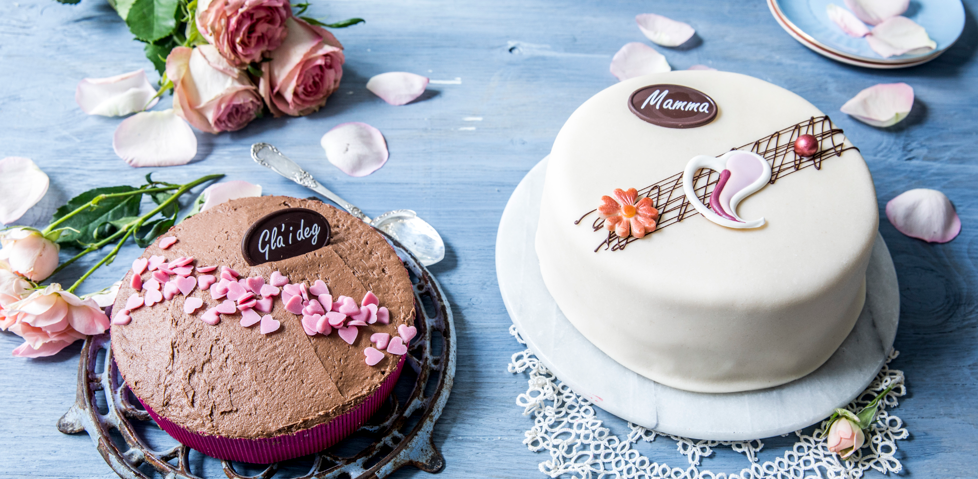 To kaker, en sjokoladekake med rosa blomster og en hvit marsipankake med blomsterdekorasjon, omgitt av rosa roser og blomsterblad på et blått bord, som kan være til morsdag.