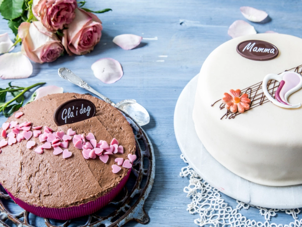 To kaker, en sjokoladekake med rosa blomster og en hvit marsipankake med blomsterdekorasjon, omgitt av rosa roser og blomsterblad på et blått bord, som kan være til morsdag.