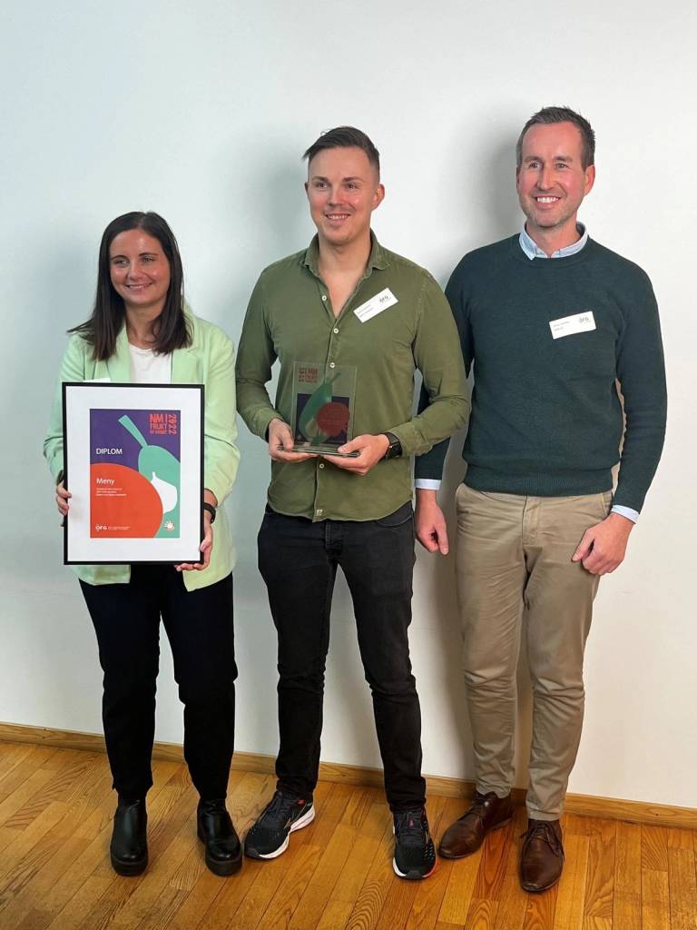 Tre personer står side om side, og den midterste holder en pris eller utmerkelse, mens den venstre personen holder et sertifikat eller en plakat, alle med navneskilt på seg.