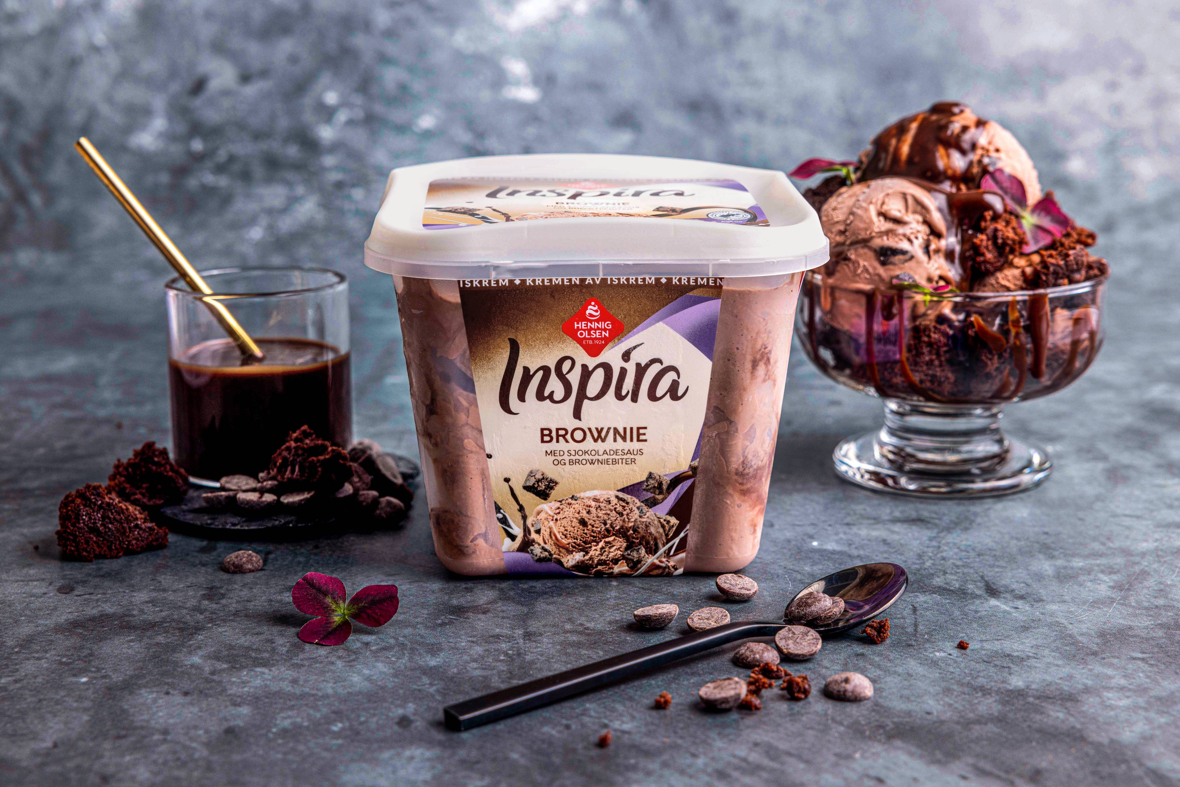 En boks med Inspira Brownies iskrem omgitt av brownies, sjokoladebiter og en kopp med sjokoladesaus, på en mørk bakgrunn.