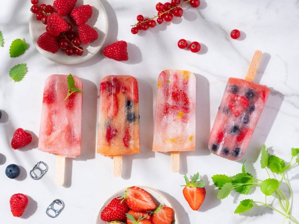 Fire ispinneformede iskrem med røde, blå og svarte bær, omgitt av frukt og bær på en hvit bakgrunn.