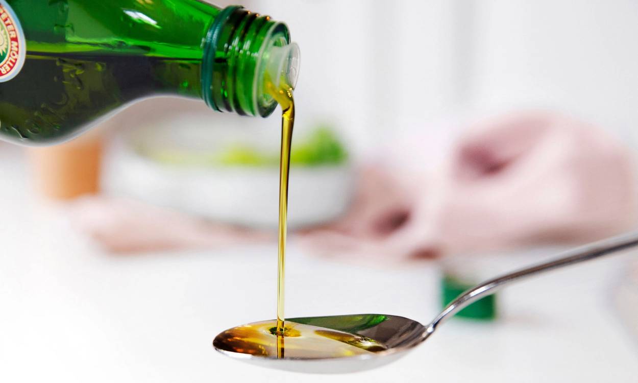 En skje som blir fylt med tran fra en grønn flaske, med en skål og et kjøkkenhåndkle i bakgrunnen.