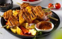 Grillspyd med svinekjøtt, paprika og ananas på en svart tallerken med saus ved siden av.