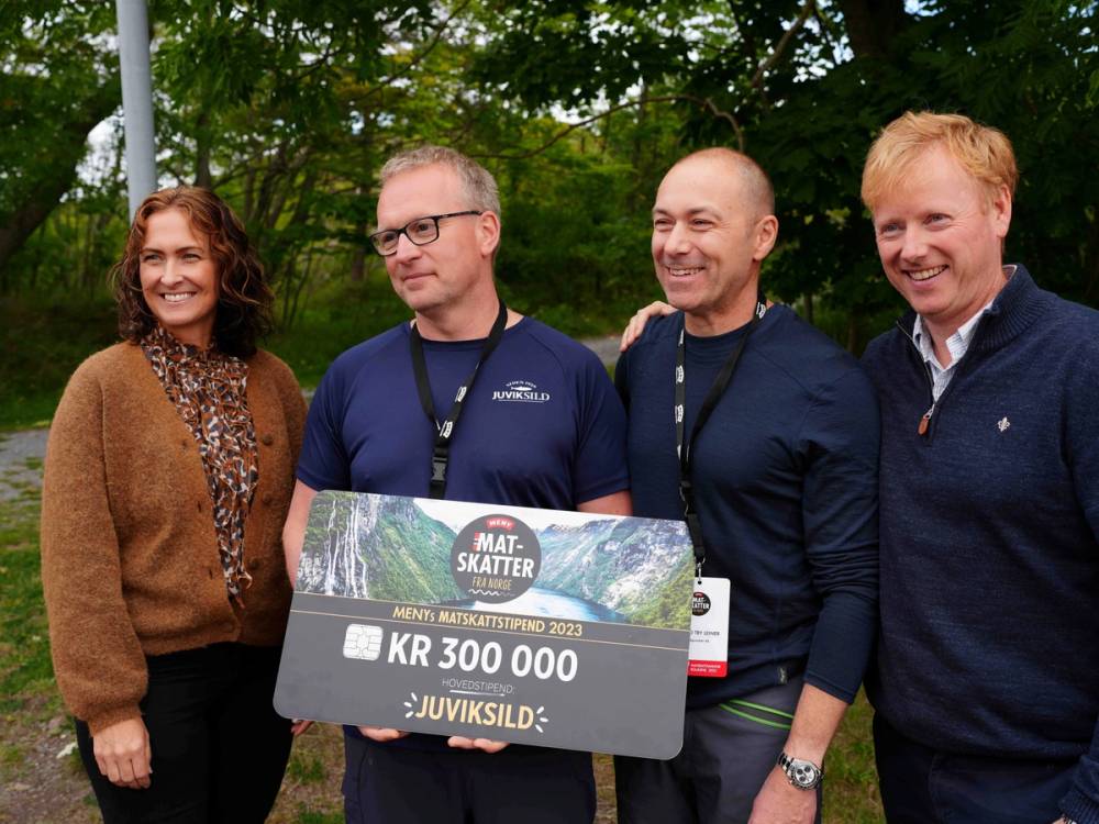 Fire personer står sammen utendørs, og en av dem holder et stort sjekk med beløpet 300 000 kroner, med skog i bakgrunnen.