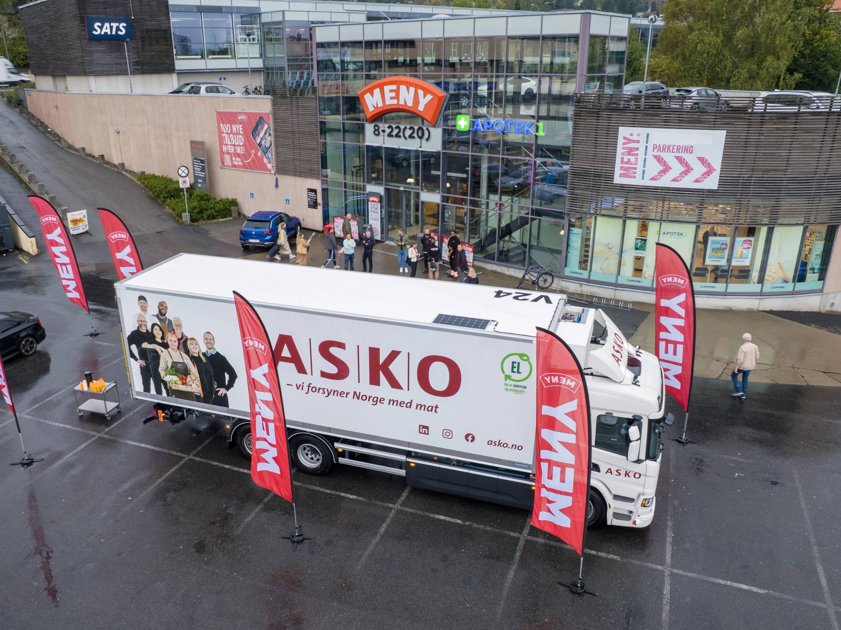 En stor hvit lastebil med ASKO-logo står parkert foran en bygning, omgitt av flere røde flagg med WEKY-tekst, og det er folk som går i området.