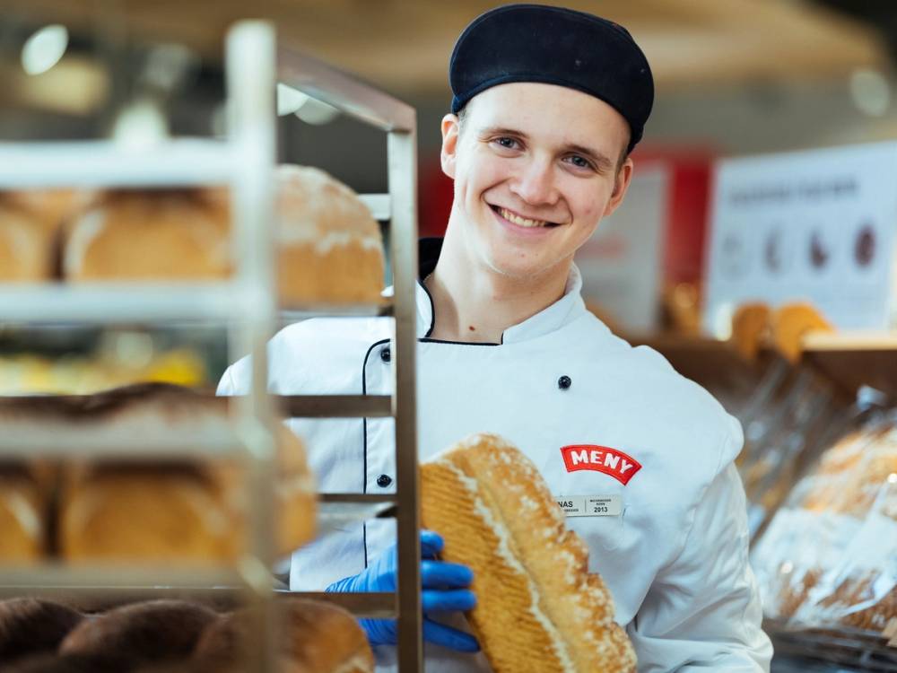 En baker i en butikk holder en skive brød, omgitt av ulike typer brød og bakervarer.