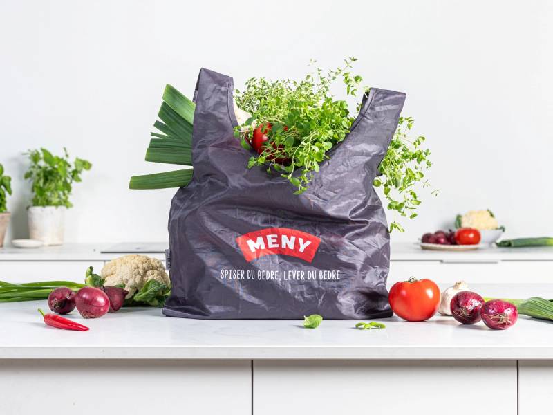 En gjenbrukspose fylt med grønnsaker og urter på kjøkkenbenk, omgitt av flere grønnsaker og urter.