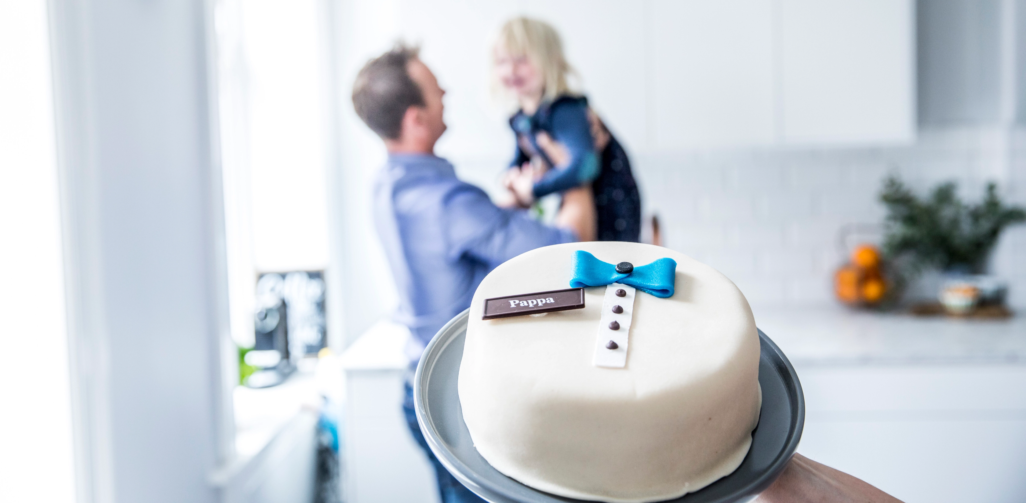 En person holder en hvit bursdagskake med blå dekorasjon i forgrunnen, mens en annen person smiler og snakker i bakgrunnen i et kjøkken.