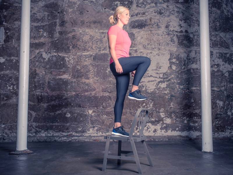 En kvinne står på en stol med ett ben hevet, mens hun holder balansen i en treningsøvelse kalt kassesteg, i et treningsrom med en murvegg i bakgrunnen.