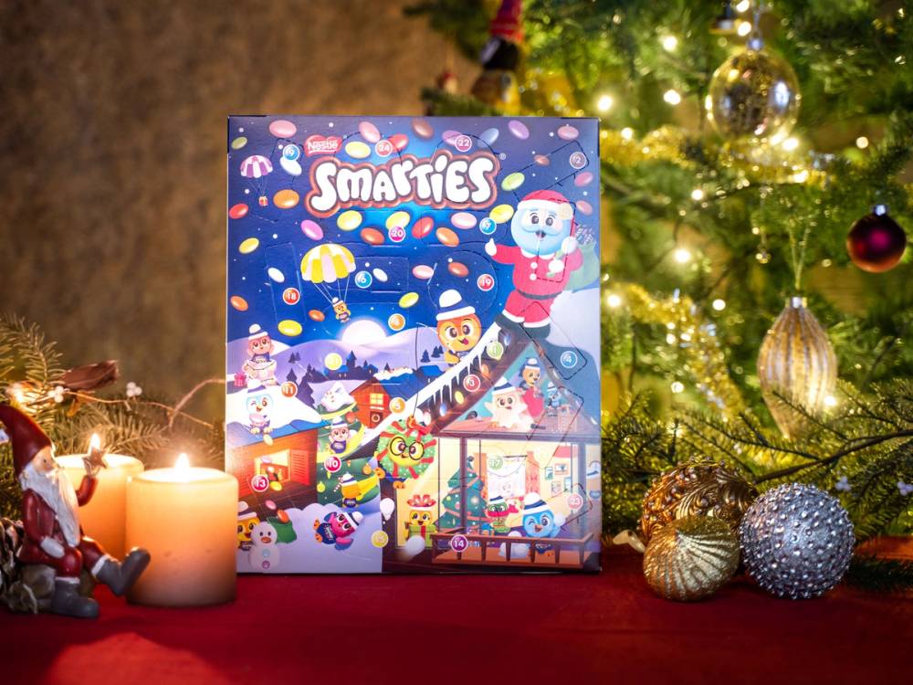 En Smarties-kalender med juletema står foran en julegran med pynt og lys, omgitt av juledekorasjoner og lyslenker.