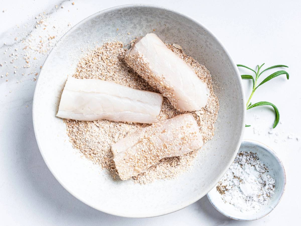 Fiskepinner dekket med mel og brødsmuler på en hvit tallerken, klar til å bli stekt eller bakt.