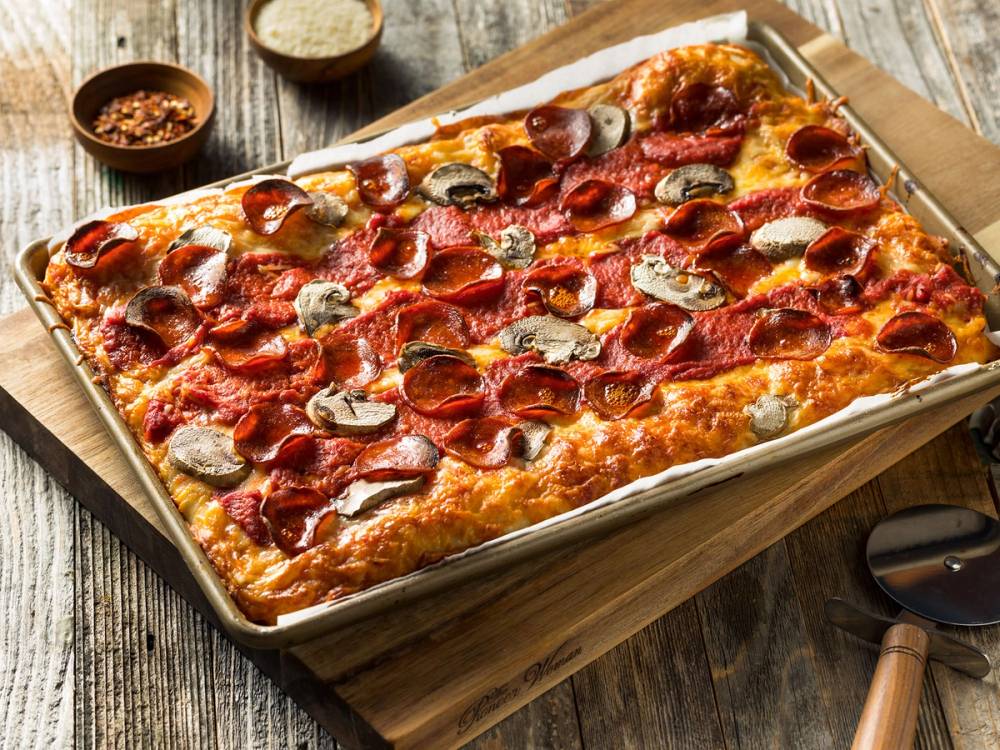 En stor Detroit-style pizza med pepperoni, sopp og ost på en bakplate, omgitt av små skåler med krydder og en tresleiv.