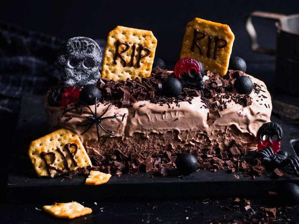 En Halloween-inspirert sjokoladekake pyntet med kjeks, bær og små skjeletter, med mørk bakgrunn og skumle elementer.
