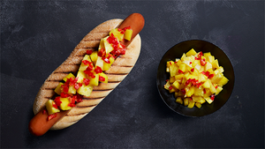 En pølse i brød med mango- og paprika-salat på siden, servert på en mørk bakgrunn.