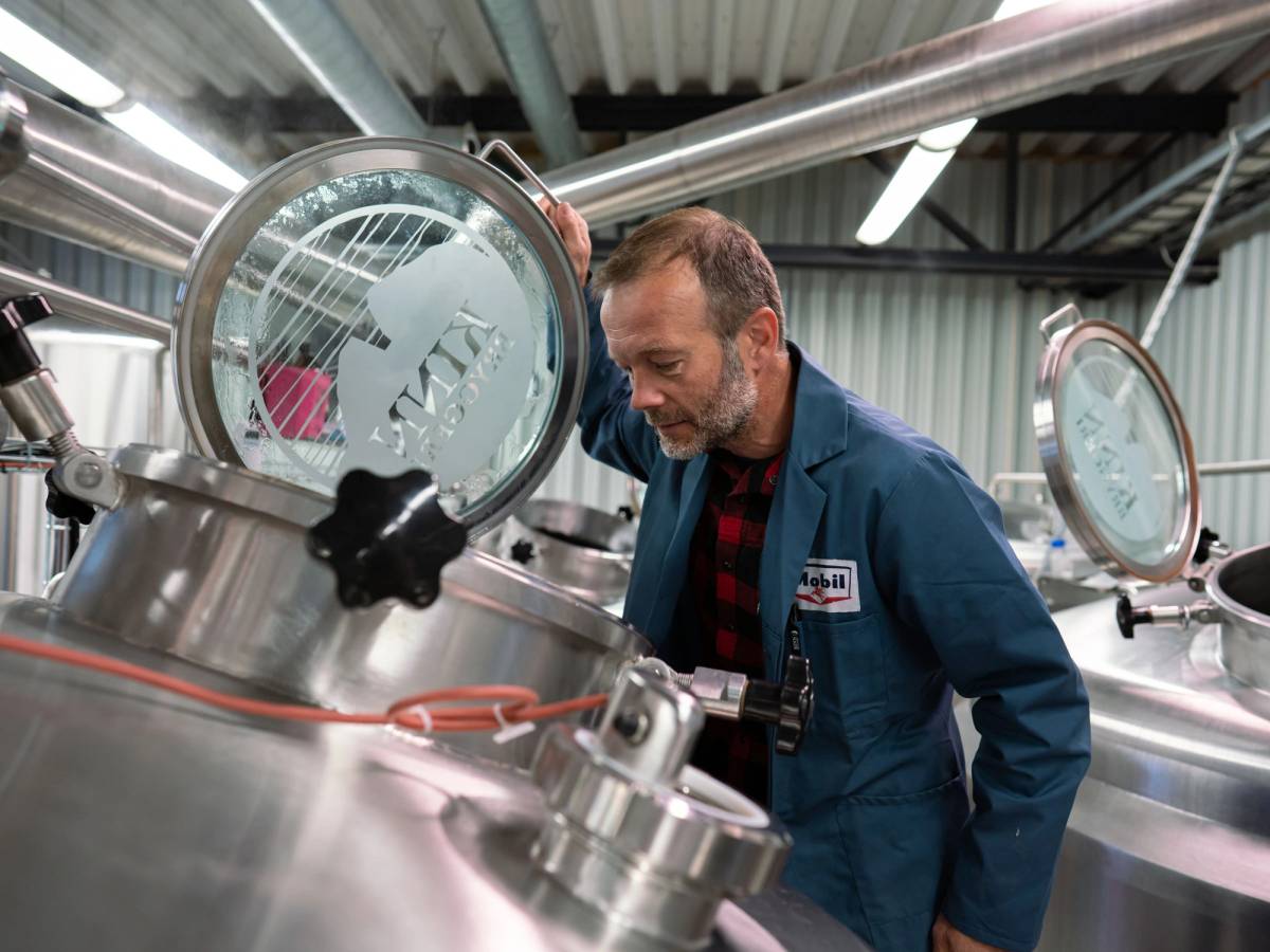 En person i blå arbeidsdress undersøker en stor tank på et industrielt anlegg, med fokus på tankens inspeksjonsluke.