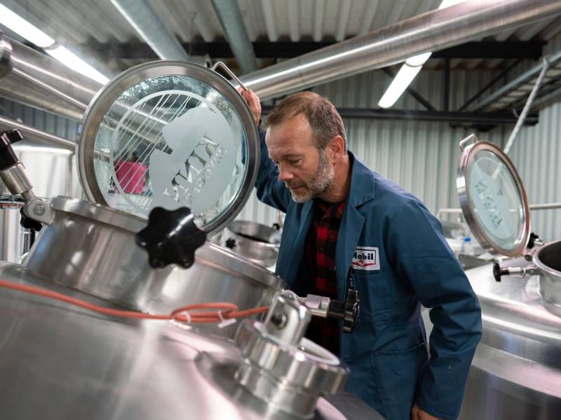 En person i blå arbeidsdress undersøker en stor tank på et industrielt anlegg, med fokus på tankens inspeksjonsluke.