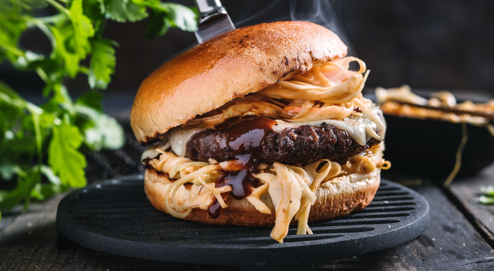 En saftig burger med kjøttkake, ost, løk og saus i et mykt hamburgerbrød, plassert på en svart grillrist med grønne urter i bakgrunnen.