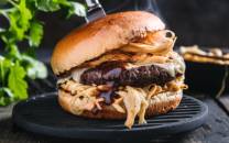 En saftig burger med kjøttkake, ost, løk og saus i et mykt hamburgerbrød, plassert på en svart grillrist med grønne urter i bakgrunnen.