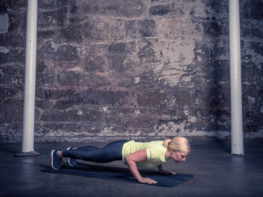 En kvinne gjør push-ups på en treningsmatte i et treningsstudio med murvegg i bakgrunnen.