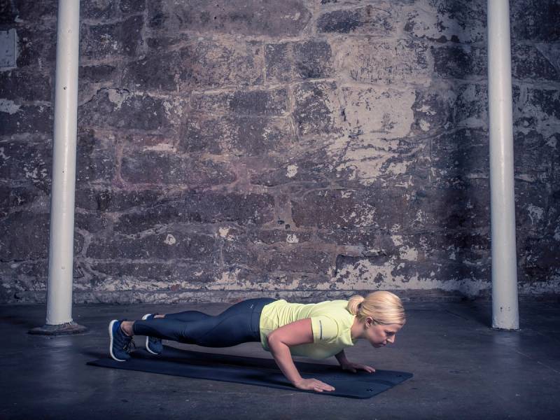 En kvinne gjør push-ups på en treningsmatte i et treningsstudio med murvegg i bakgrunnen.
