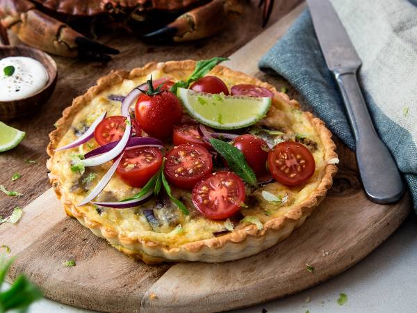 Krabbeterte med tomatsalat, pyntet med cherrytomater, løk og lime, på et rundt trefat med en kniv og en skje ved siden av.