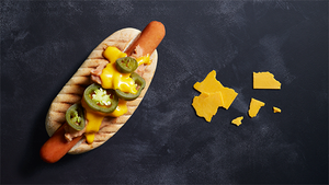 En hotdog med pølse, gul sennep, syltede agurkskiver og løk på et oval fat, plassert på en svart bakgrunn med en gul kartskisse av Texas til høyre.