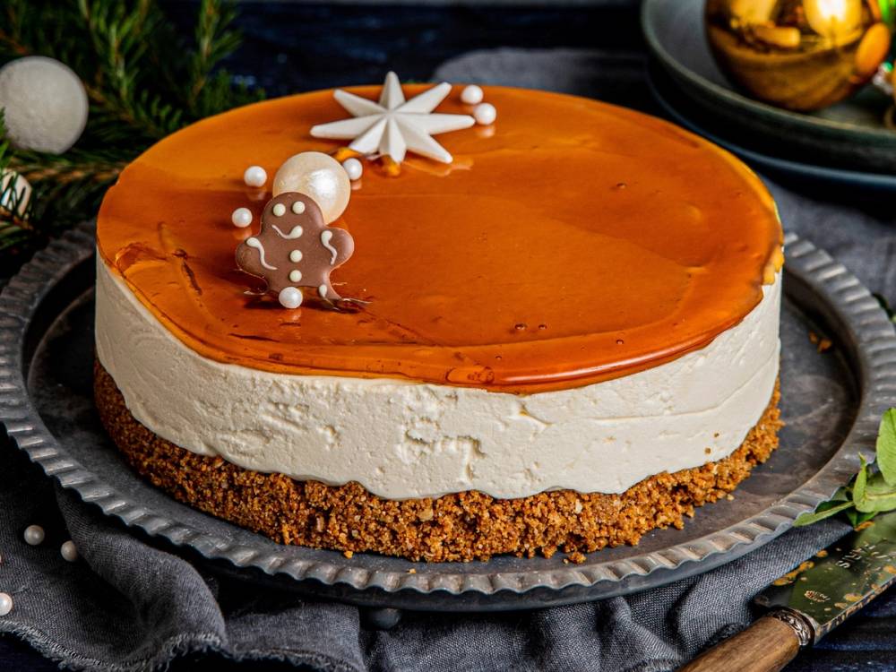 En ostekake med en karamellfarget topp, pyntet med små hvite kuler og en liten pepperkakefigur, plassert på en sølvfat med grønt dekor rundt.