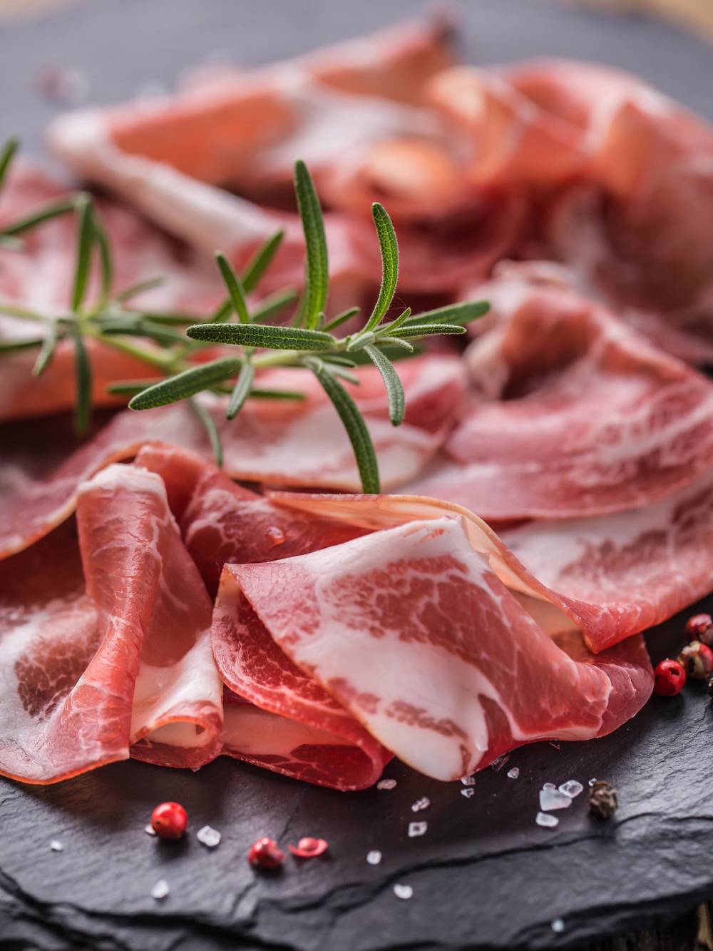 Spekeskinke høykant med fersk rosmarin og pepperkorn på en svart steinplate.