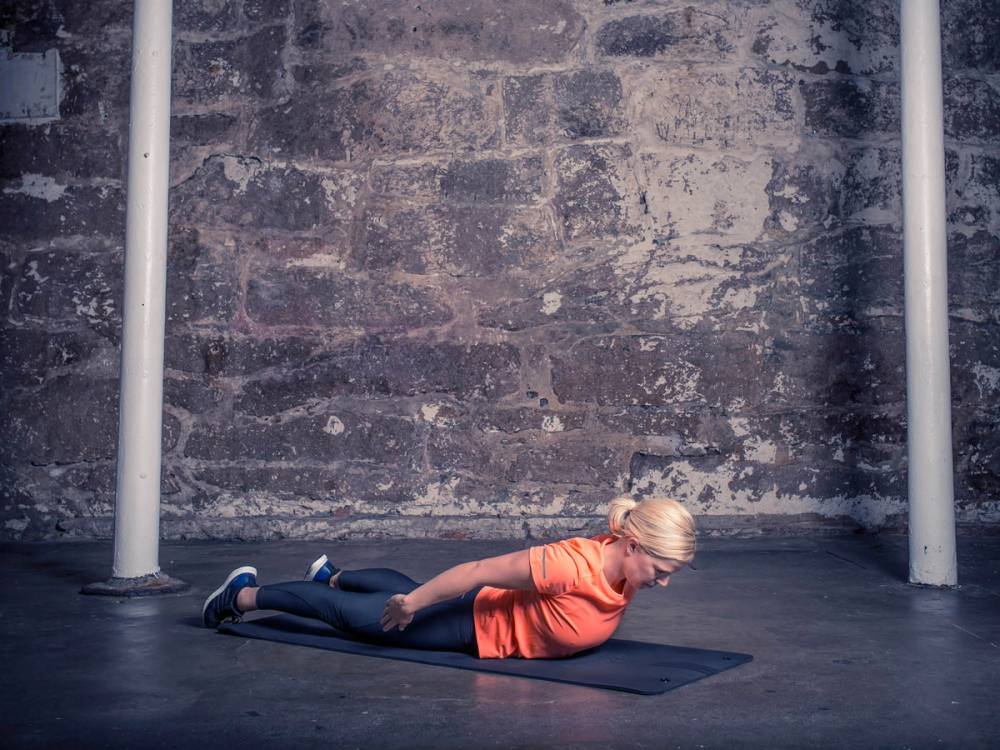 En kvinne gjør rygghev på en treningsmatte i et treningsrom med murvegg og stenger på sidene.