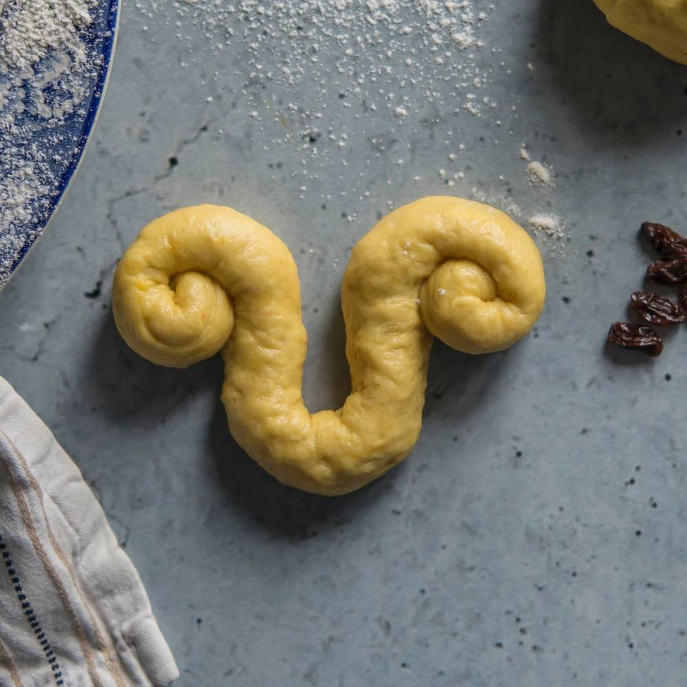 En lussekatt formet som en lilje på en kjøkkenbenk med mel og småkaker rundt.