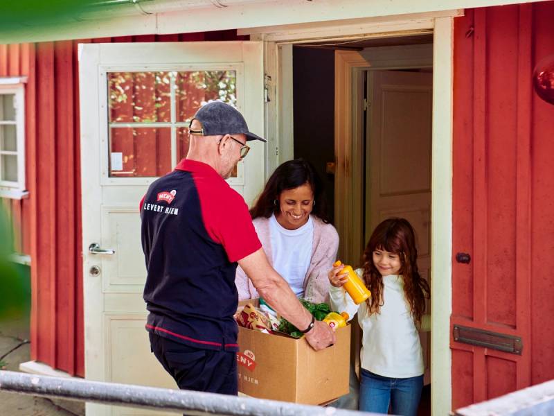 En mann i en meny-levert uniform deler ut mat til to unge jenter utenfor en rød husdør.