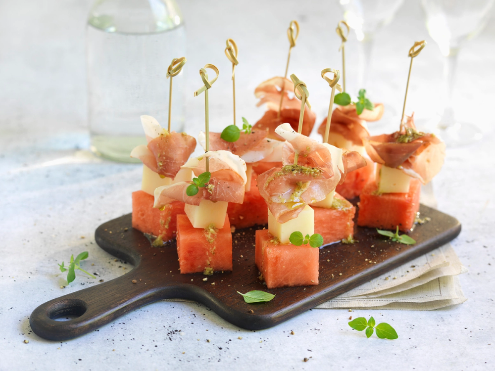 Melonspyd med spekeskinke, manchego-ost og små grønne urter på en svart serveringsplate