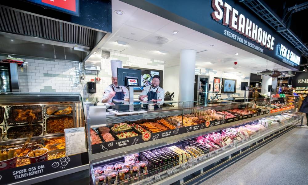 To ansatte som selger fersk kjøtt i en kjøttdisk i en matbutikk, med stearhouse-skilt i bakgrunnen.