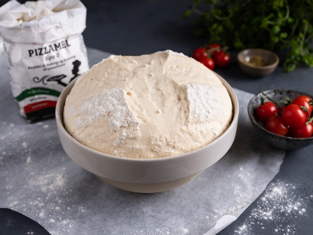 Pizzadeig i en stor bol med ingredienser og tomater rundt på et kjøkkenbord.