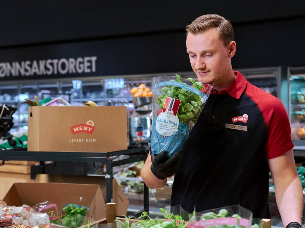 En ansatt i Meny-butikk holder en pose med grønnsaker foran en frukt- og grønnsaksavdeling.