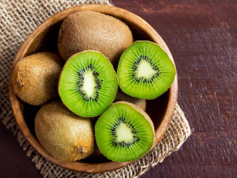 En skål med hele kiwi og skivede kiwi med grønn fruktkjøtt og svarte frø, plassert på en trefjøl.