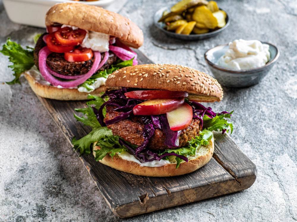 To veganske burgere med grønnsaker og saus på et trebrett, omgitt av små skåler med pickles og dressing.